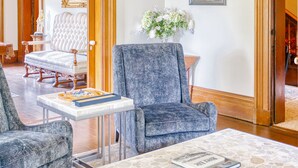 Amplia sala de estar con una lámpara de araña, una butaca blanca tapizada, un sillón azul y una mesa de centro con libros.