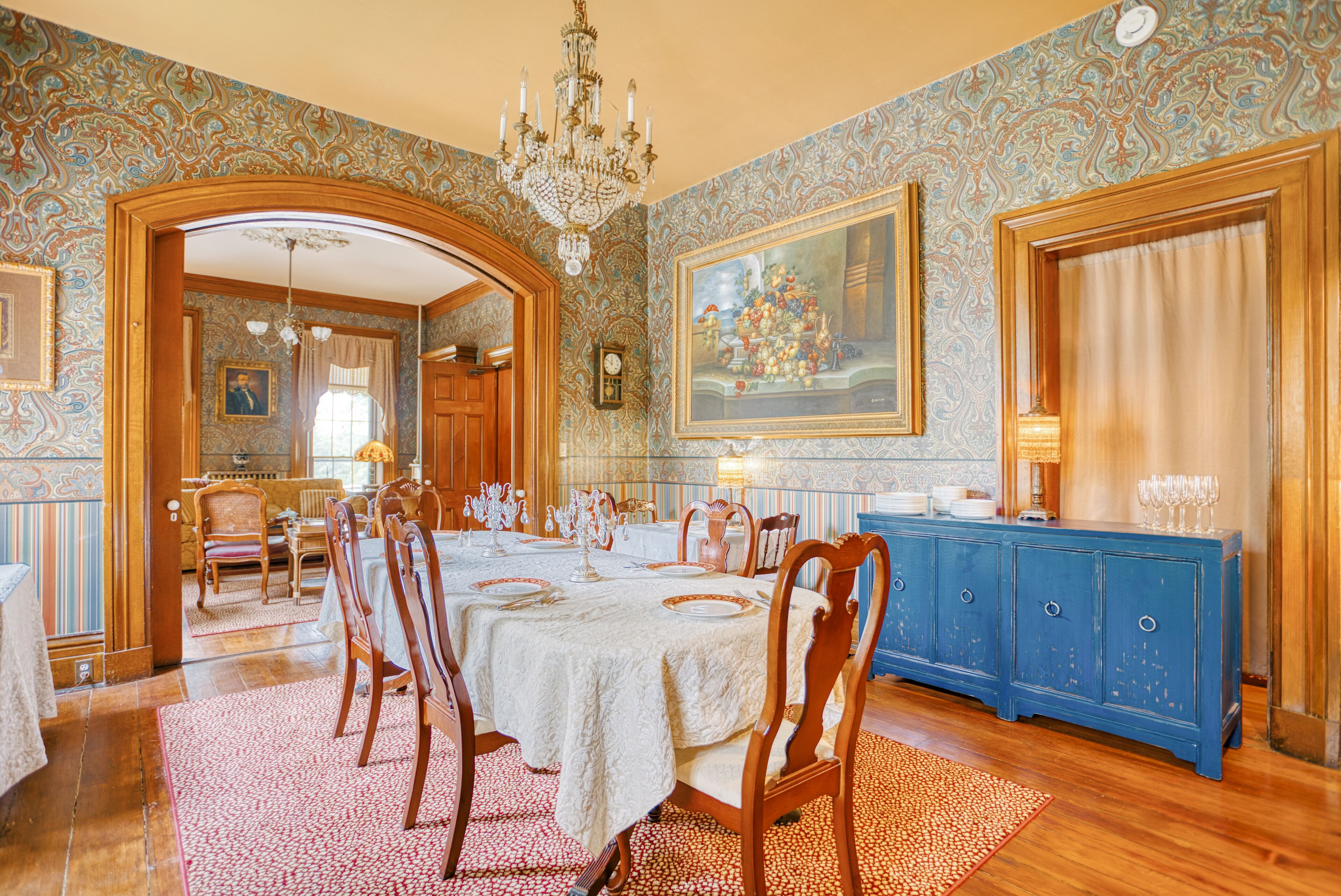 A dining room with a long table set for six, surrounded by wooden chairs. A blue cabinet with glassware is against the wall.