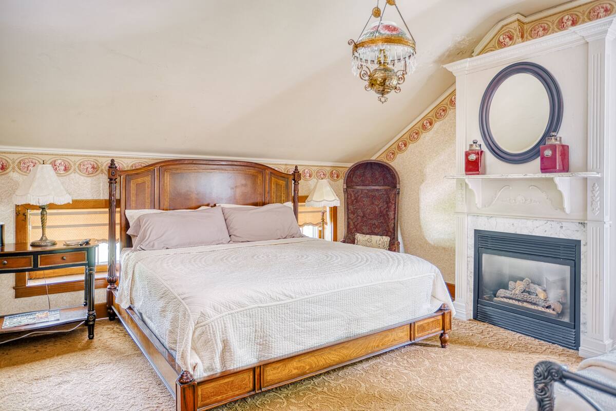 Ein Schlafzimmer mit einem hölzernen Bett, einem Nachttisch, einer Lampe, einem Kamin und einem Spiegel darüber.
