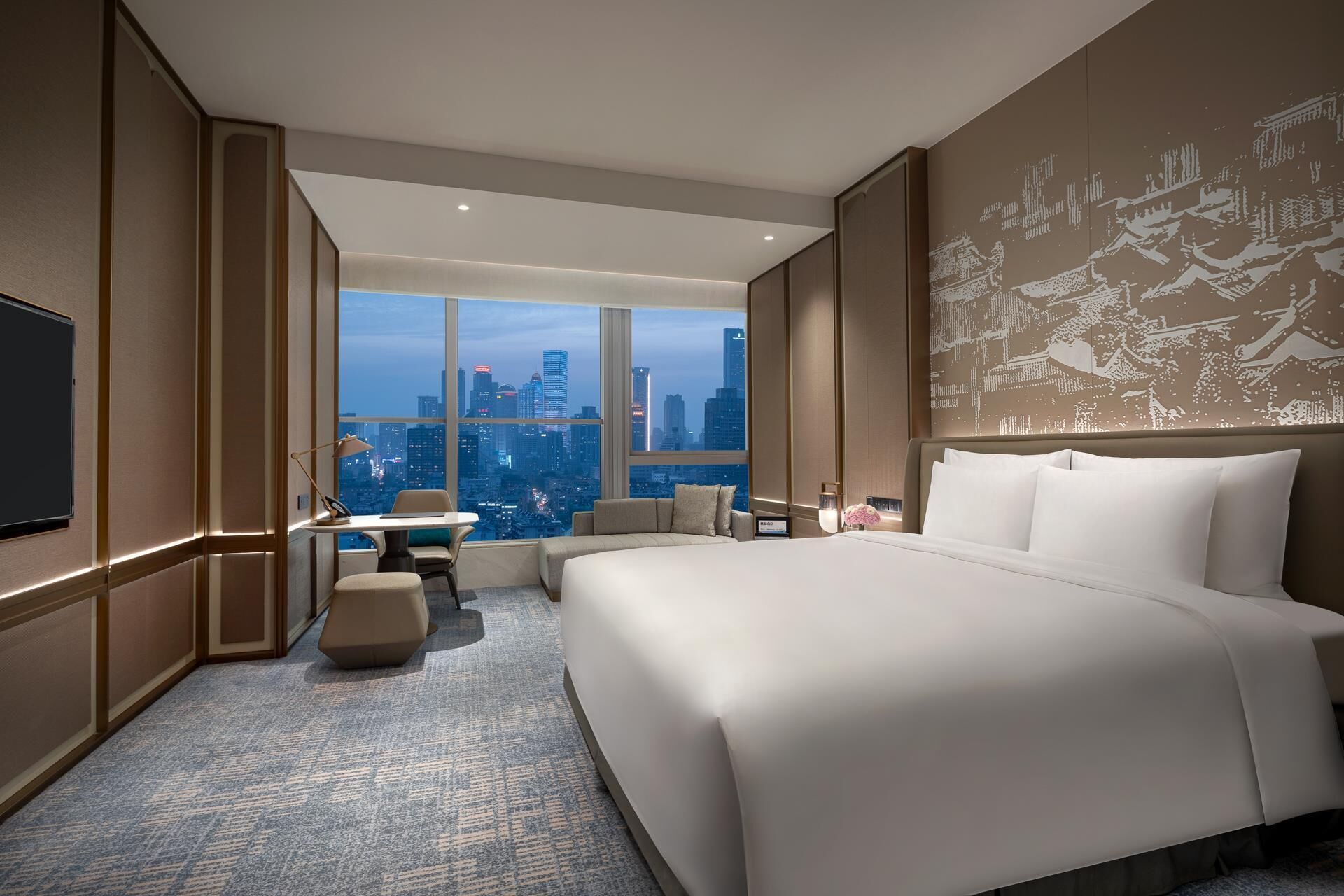 A modern hotel room with a large bed, a desk, a chair, and a view of a city skyline.