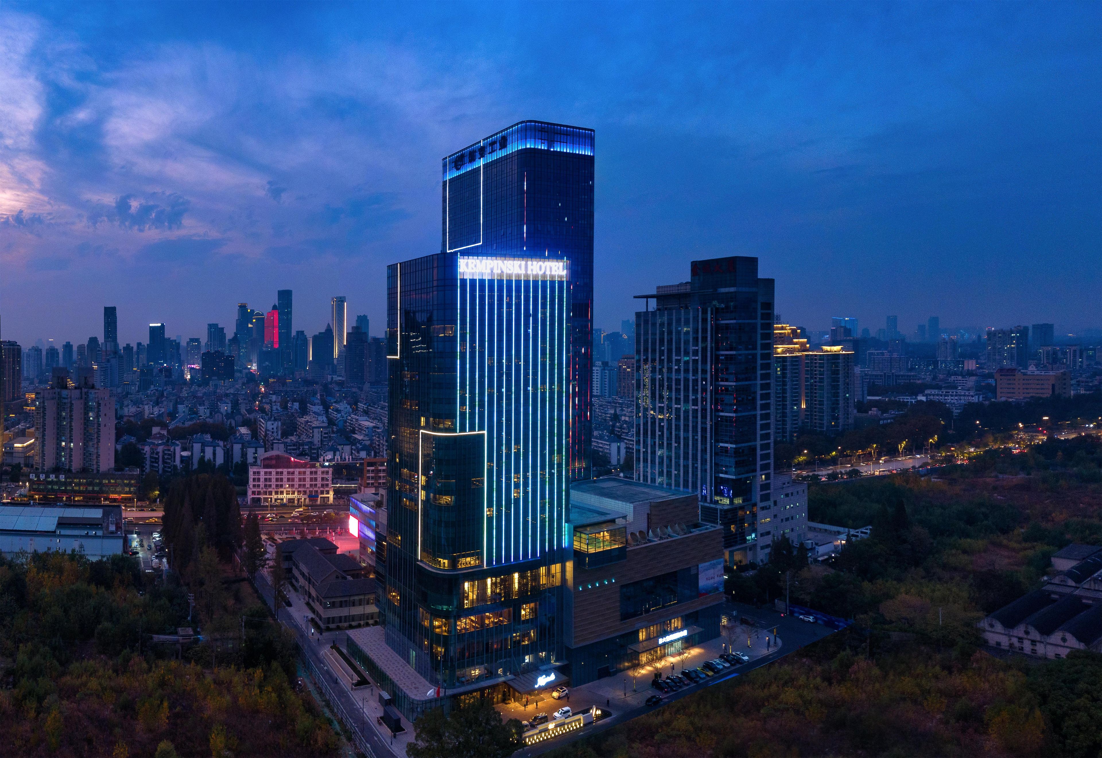 Un hôtel de grande hauteur, doté d’une façade moderne en verre et d’une enseigne lumineuse, s’élève au milieu d’un paysage urbain au crépuscule.