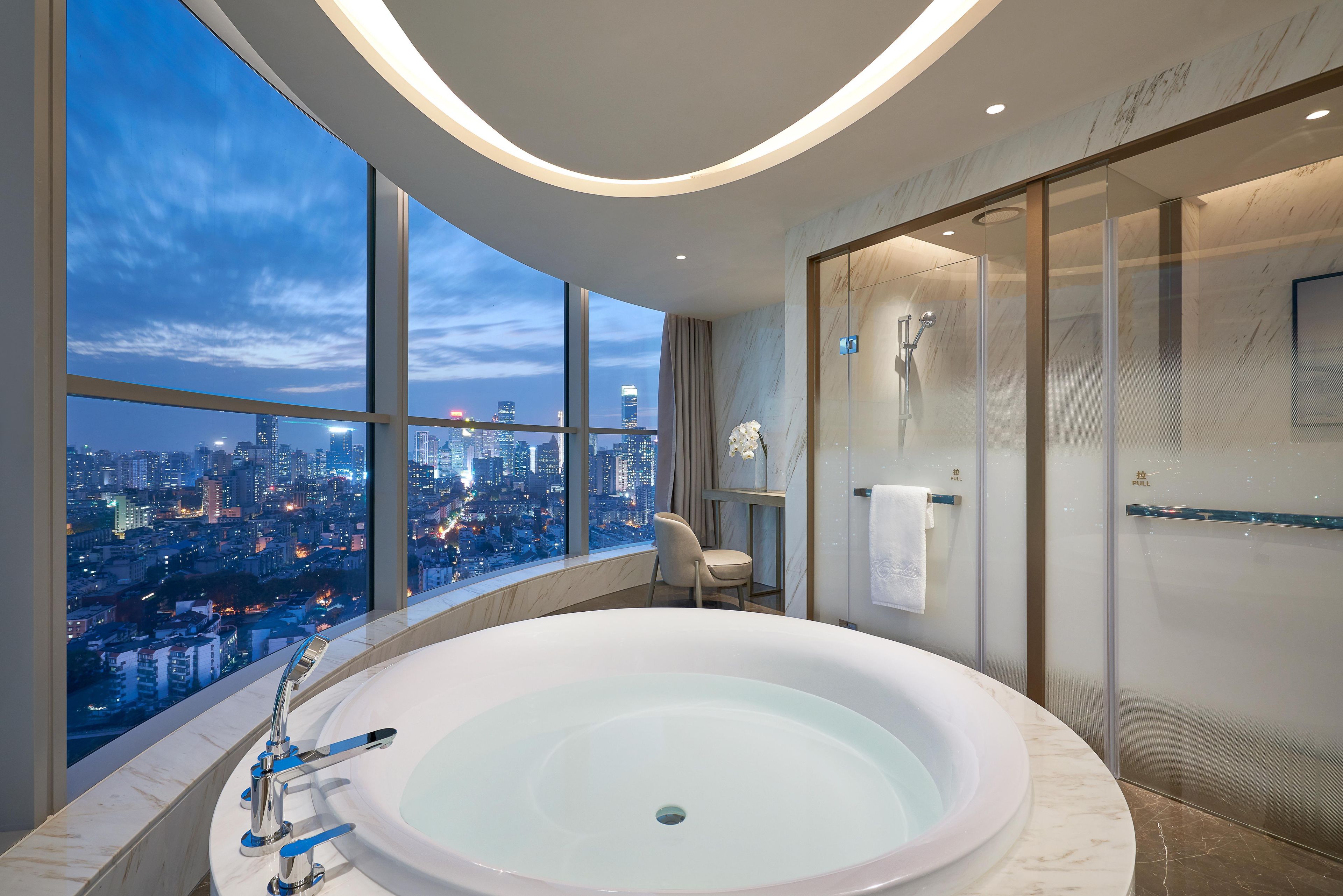A modern bathroom with a large bathtub, a glass-enclosed shower, and a city view.