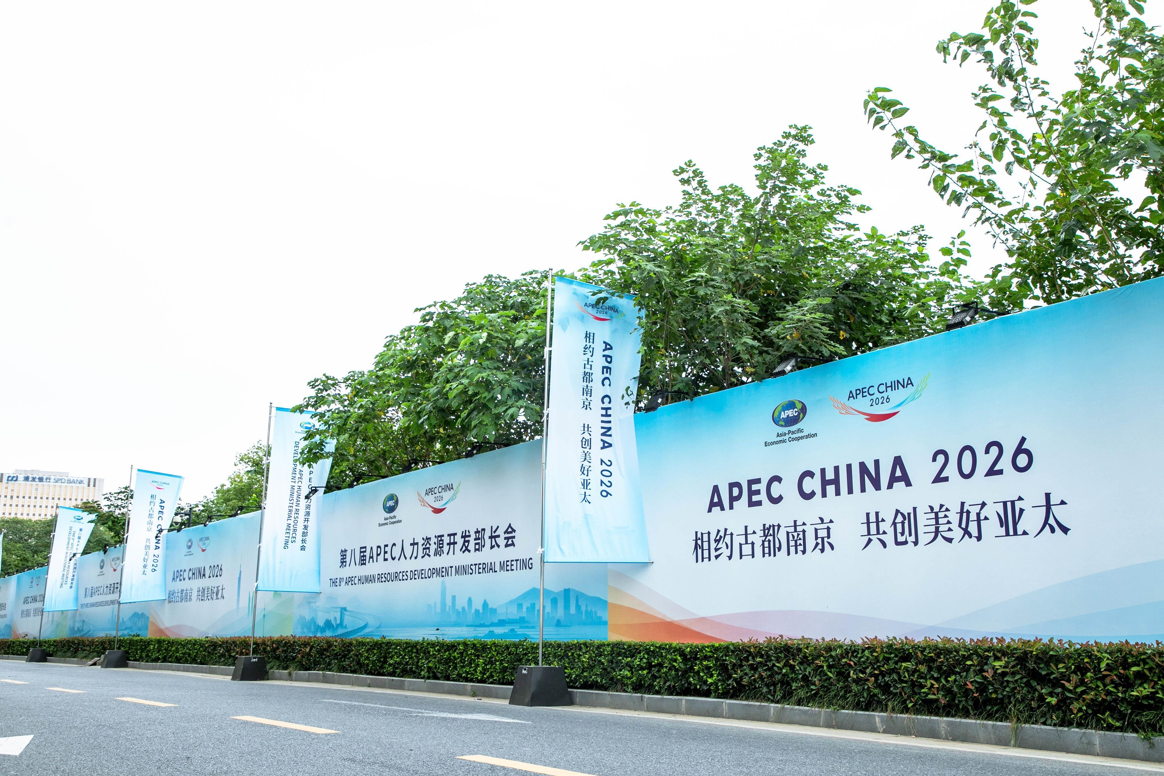 Ein großes blaues Banner mit dem Text APEC China 2026 und Logos, das von Bäumen und einer Straße gesäumt wird.