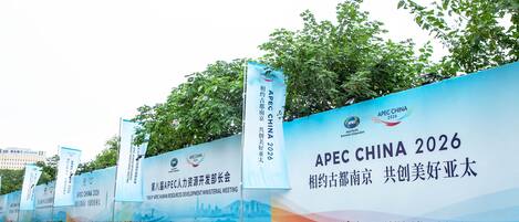 A large blue banner with APEC China 2026 text and logos, flanked by trees and a road.