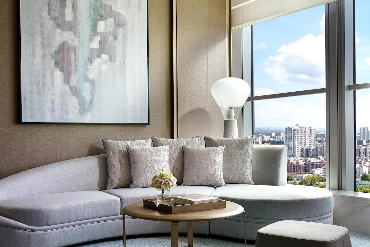 Un salon moderne avec un canapé d’angle gris, une table basse ronde et une vue sur la ville grâce à de grandes fenêtres.