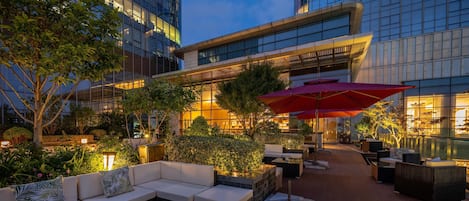 Un patio al aire libre con mesas y sillas, una piscina y un edificio con ventanas iluminadas al fondo.