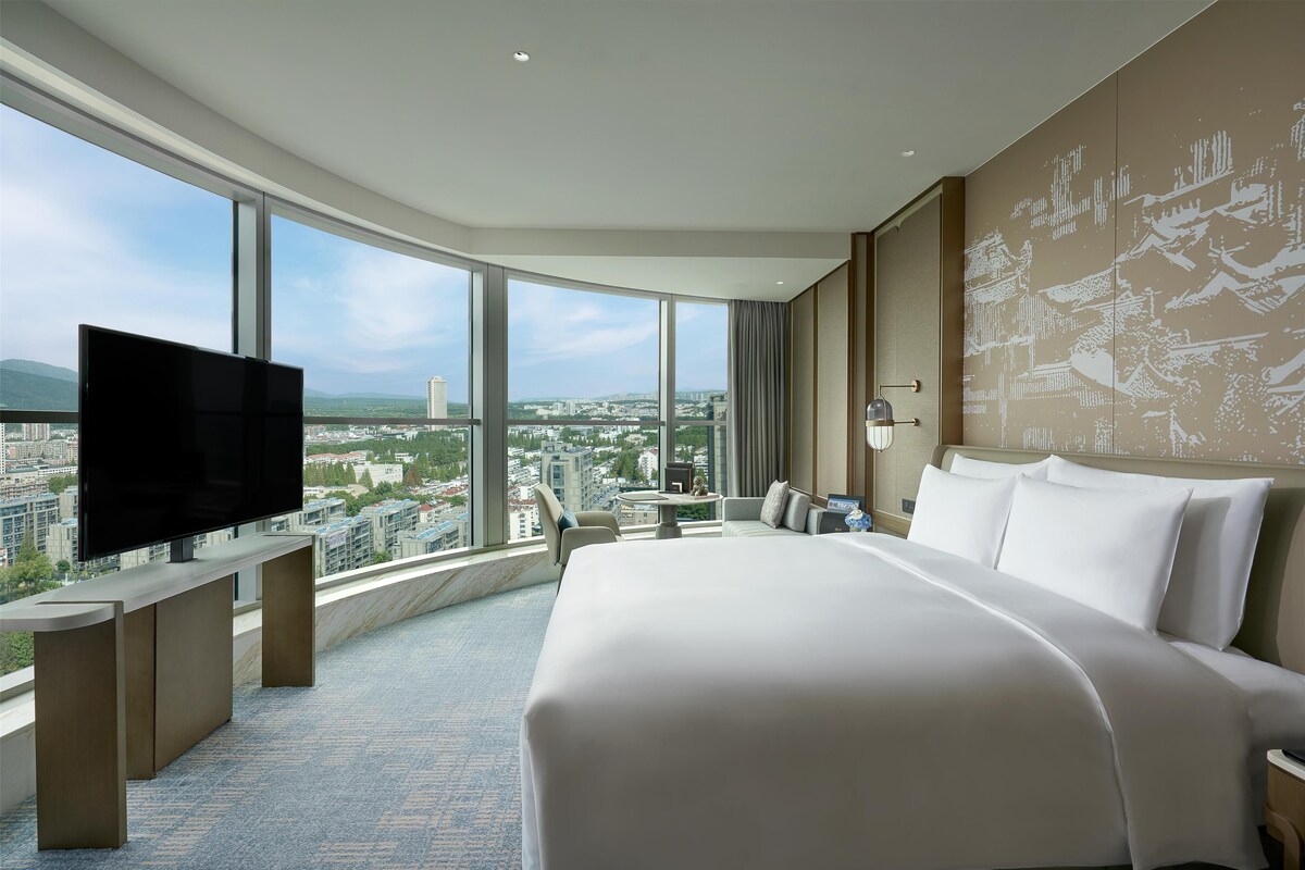 Une chambre d’hôtel moderne dotée d’un grand lit, d’une télévision à écran plat, d’un bureau et offrant une vue sur la ville grâce à de grandes fenêtres.