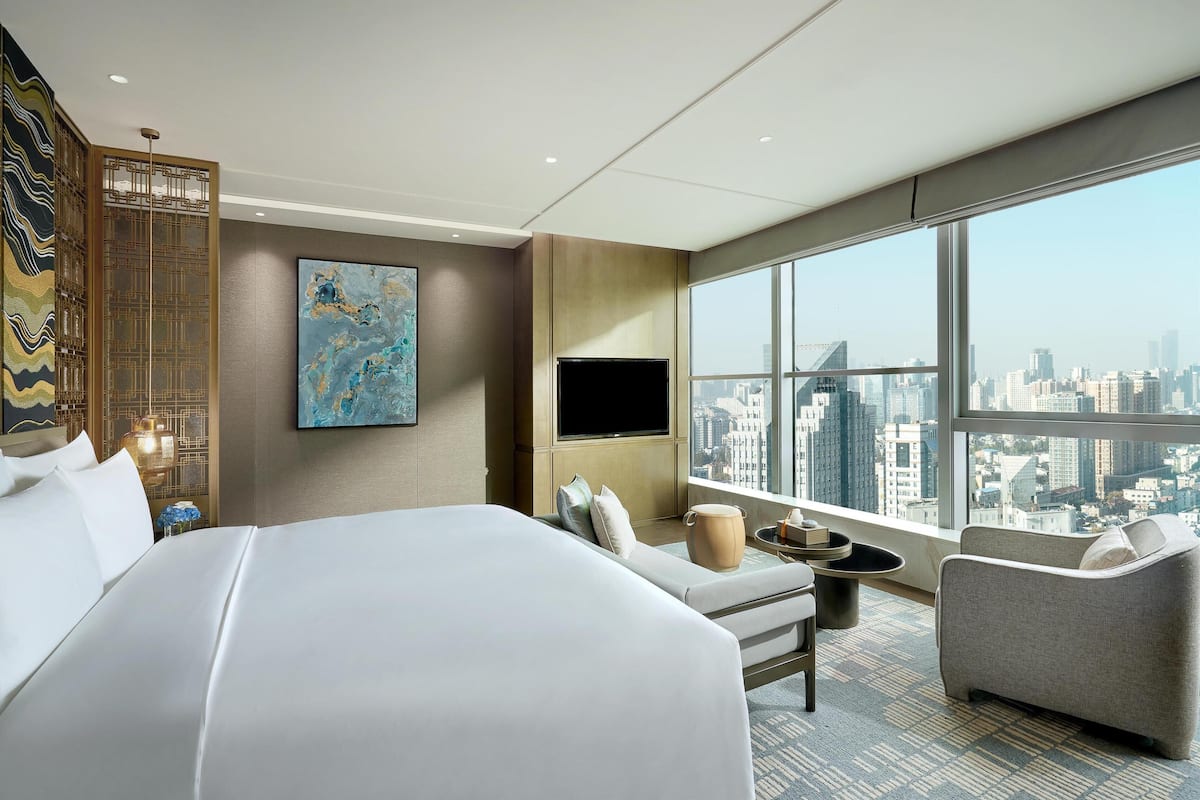 Une chambre d’hôtel avec un grand lit, un téléviseur à écran plat, une vue sur la ville, un paravent décoratif et une petite table sur laquelle est posé un vase.
