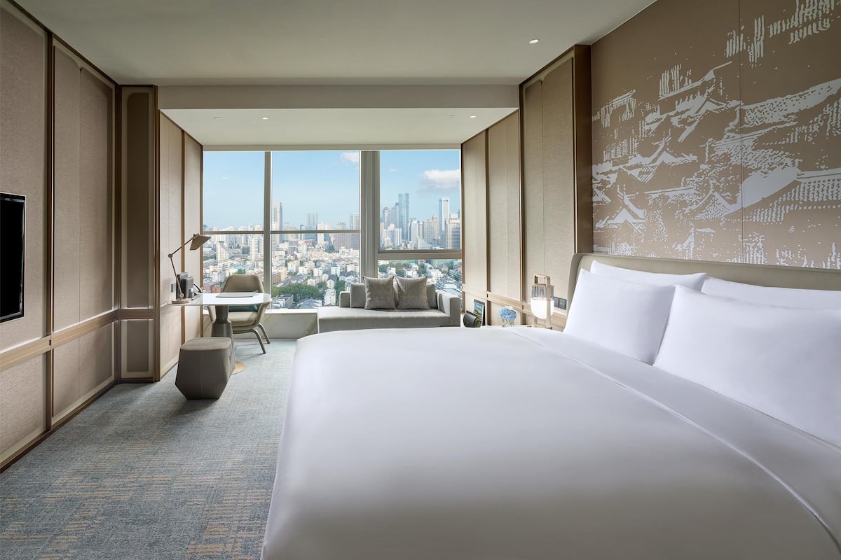 Une chambre d’hôtel avec un grand lit, un bureau, une chaise et une vue sur le paysage urbain, grâce à de grandes fenêtres.
