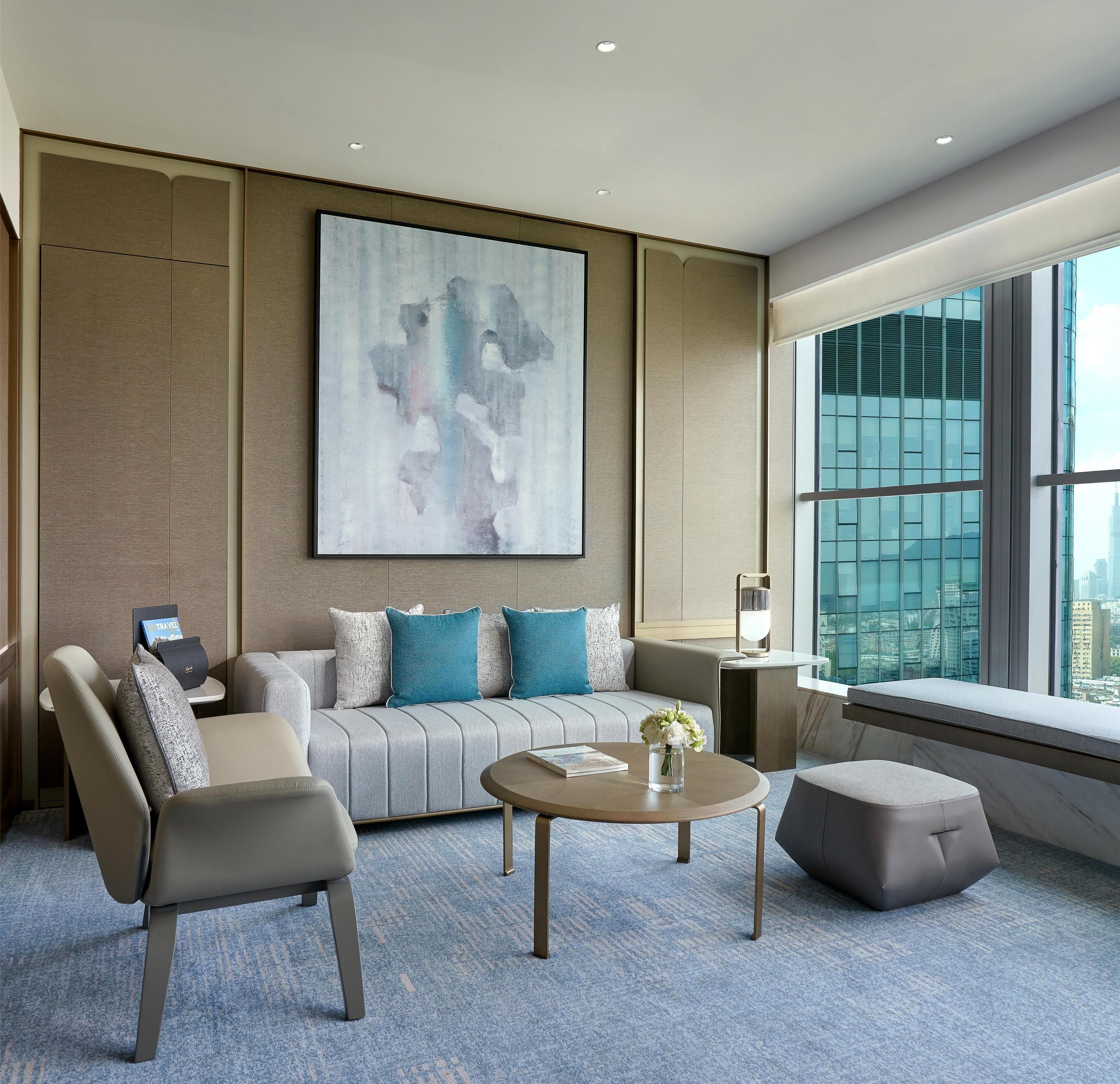 A modern hotel room with a sofa, armchair, round coffee table, and a large abstract painting on the wall.