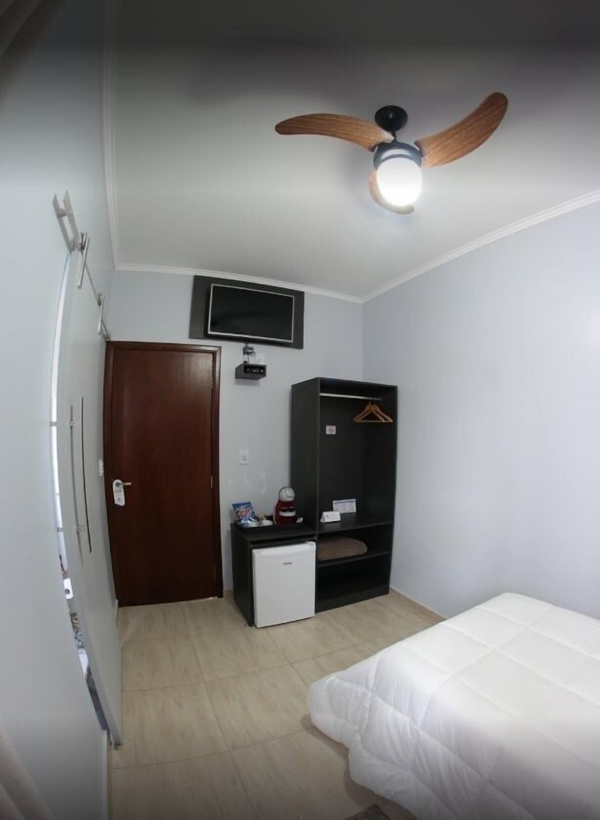 A small, clean room with a bed, a desk with a computer, a TV mounted on the wall, a wardrobe, and a ceiling fan.