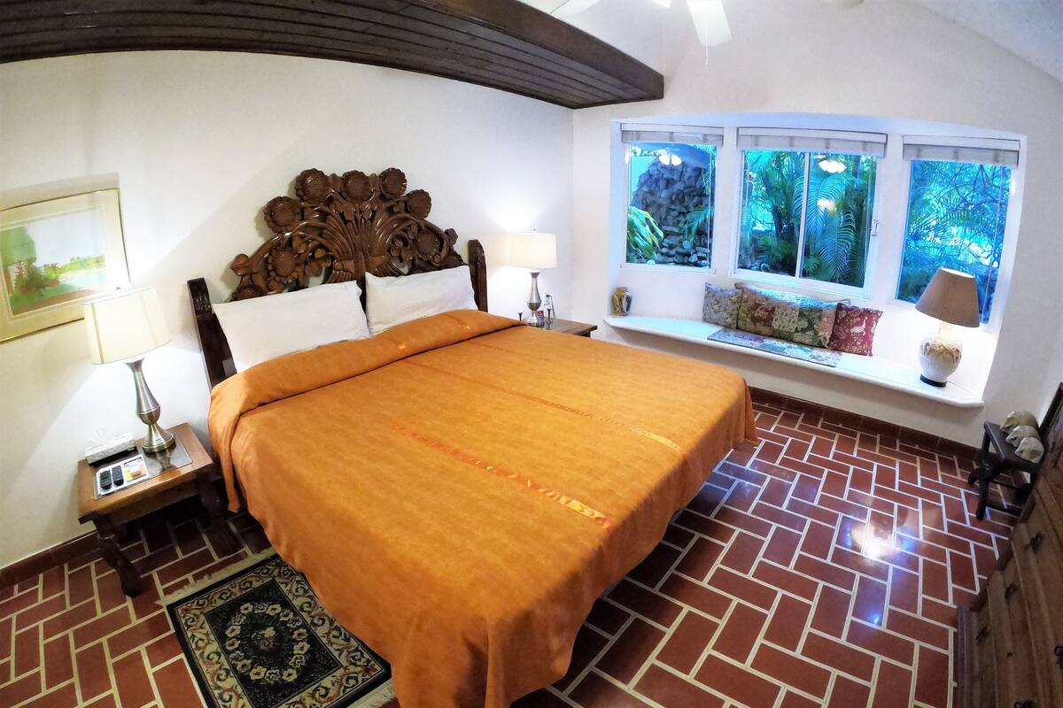 Ein Schlafzimmer mit Terrazzoboden, einem großen Bett mit orangefarbener Bettdecke, einem Holzkopfstück, einem Nachttisch mit Lampe, einem Fenster mit Blick auf Grünflächen und einer Bank mit Kissen.