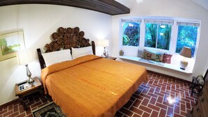 Quarto com piso de tijolos, uma cama grande com colcha laranja, cabeceira de madeira, mesa de cabeceira com abajur, janela com vista para área verde e um banco com almofadas.