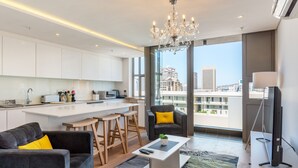 A modern living room with a sofa, coffee table, and a TV. There is a kitchen area with white cabinets and a dining table. A large window offers a view of the cityscape. - Mirage - WHosting (Cape Town)