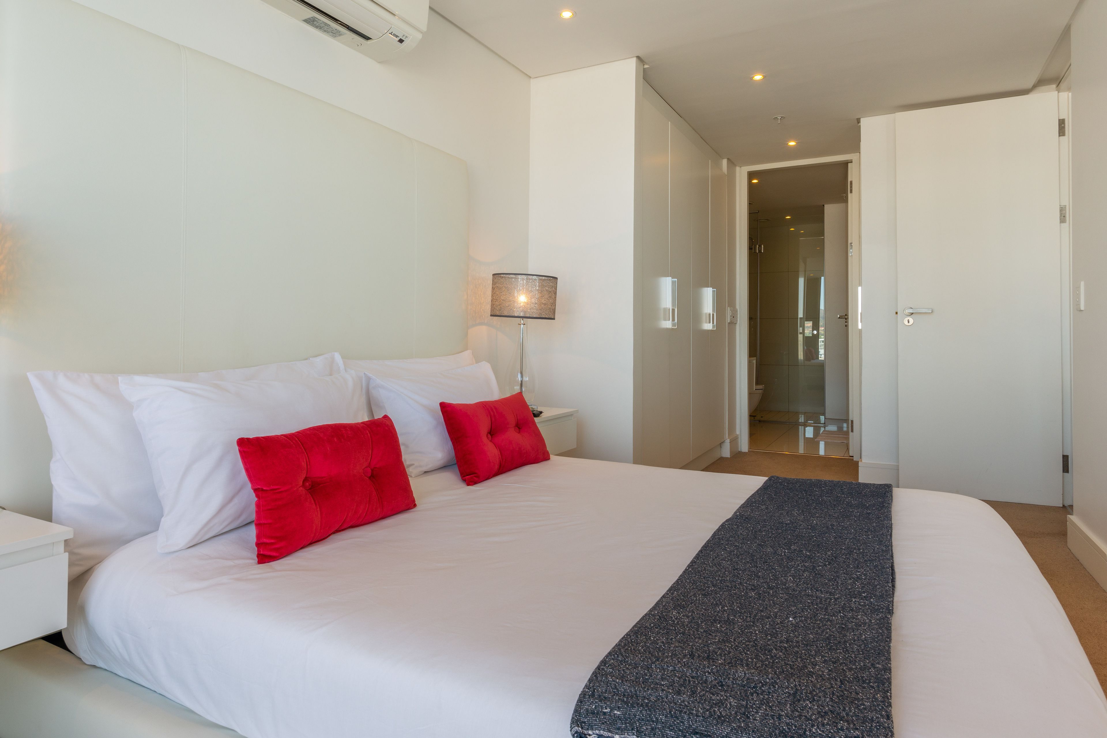 A modern hotel room with a large bed, white bedding, and red pillows. There is a bedside table with a lamp, a door leading to another room, and a view of a hallway.