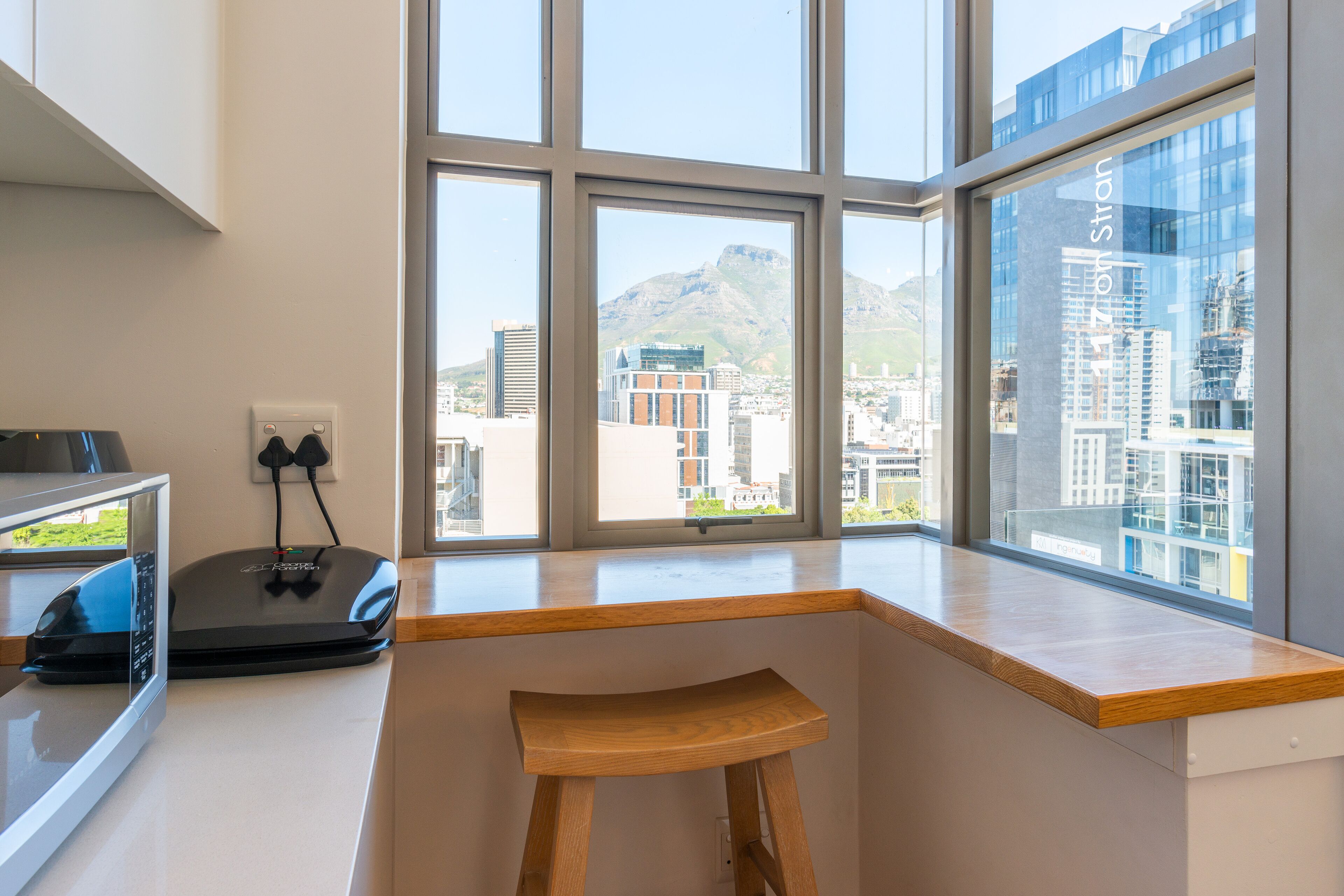 A modern kitchen with a wooden stool, a microwave, and a window with a city view.