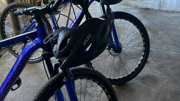 Una bicicleta azul con un casco negro y una botella de agua, estacionada en un interior con piso de concreto y vista a un parque con un banco y árboles.