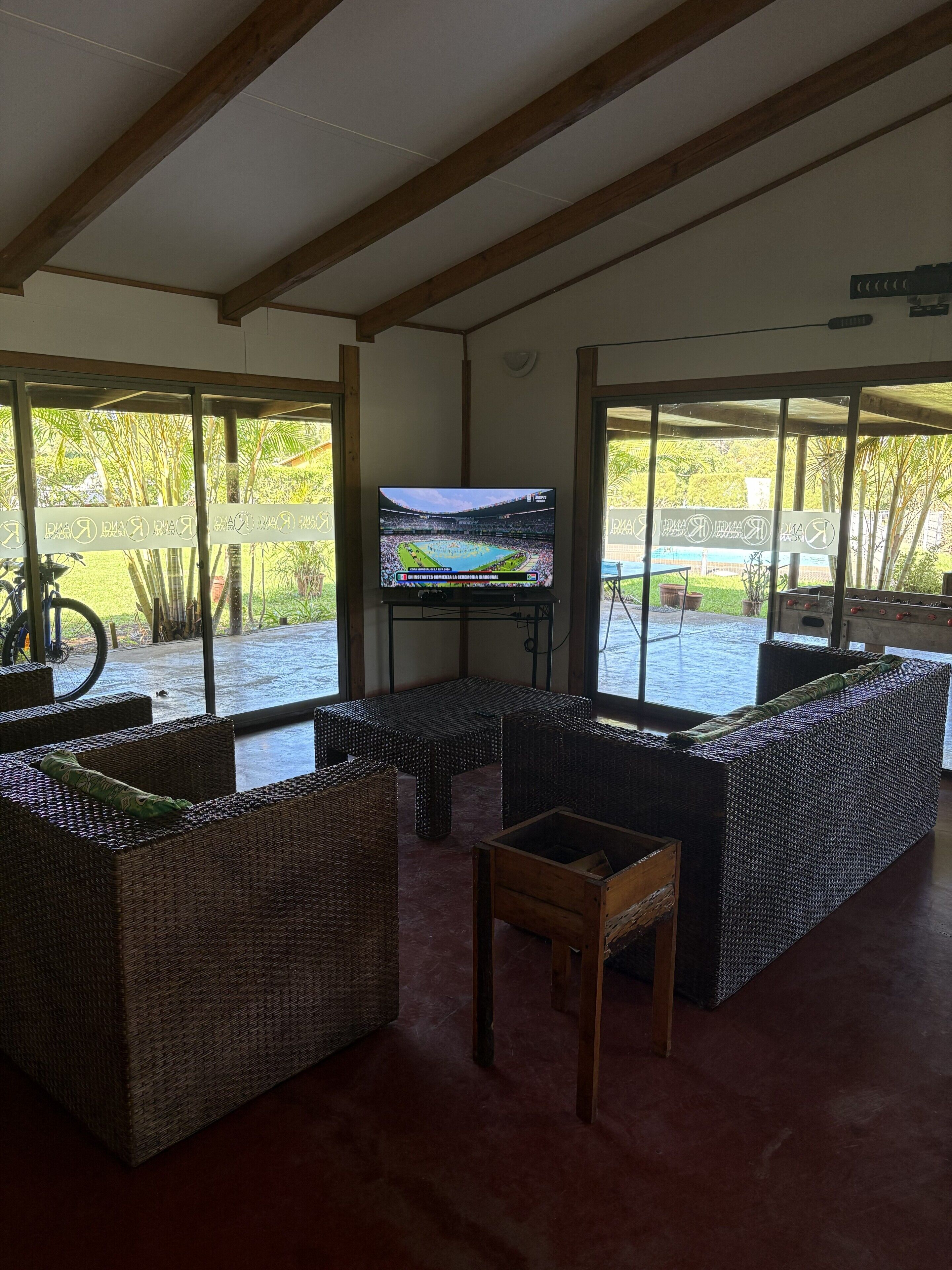 A covered outdoor seating area with wicker furniture, a television screen, and a view of a sports field.