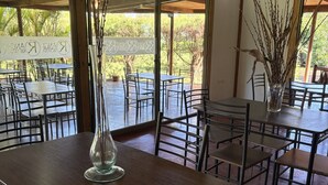 A restaurant with tables and chairs, a vase with branches, and a view of trees through large windows. - Rangi Moana (Hanga Roa)