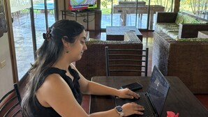 A person seated at a table with a laptop, a television in the background, and a view of the outdoors through large windows. - Rangi Moana (Hanga Roa)