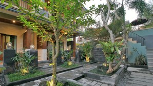 A well-maintained garden with stone pathways, various plants, and a modern building with balconies. - Kubu Rama Ubud Cottage (Ubud)