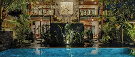 Zona de piscina con pared de piedra y escalera que conduce a un balcón, rodeada de plantas tropicales.