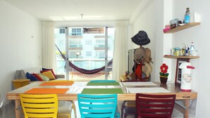 Un coin repas avec une table dressée pour quatre personnes, des chaises colorées, un hamac et une vue sur un bâtiment par la fenêtre.