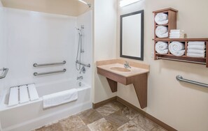 A bathroom with a bathtub, a sink, a mirror, and a shelf with towels. - My Place Hotel - Overland Park (Overland Park)