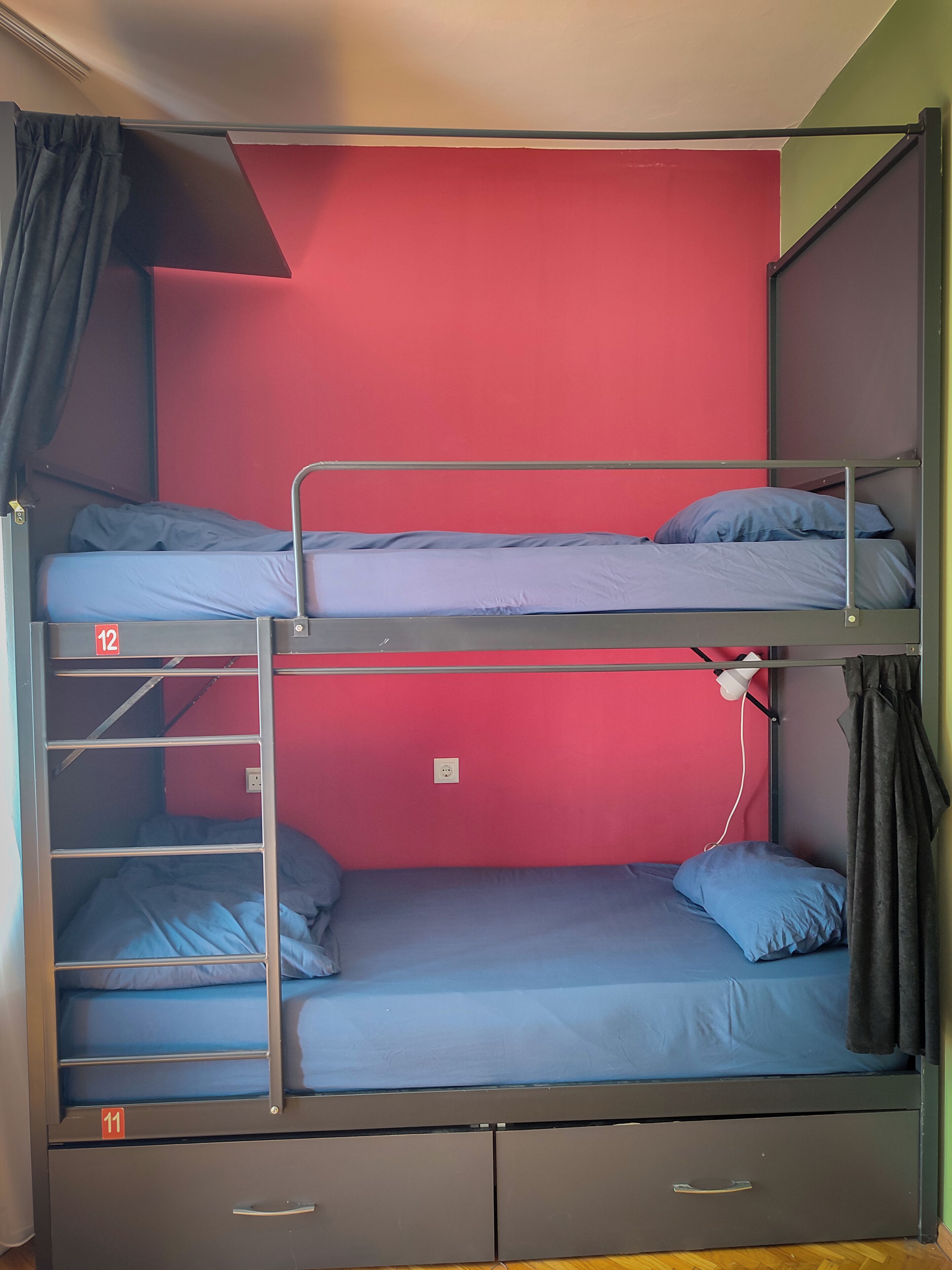 A bunk bed with three levels, each with a mattress and pillows. There is a power outlet on the wall and a curtain on the left side.