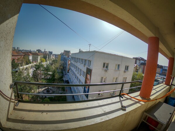A view from a balcony with a clear sky, featuring a building with multiple windows and balconies, a street below, and some greenery. - Prishtina Center Hostel (Pristina)