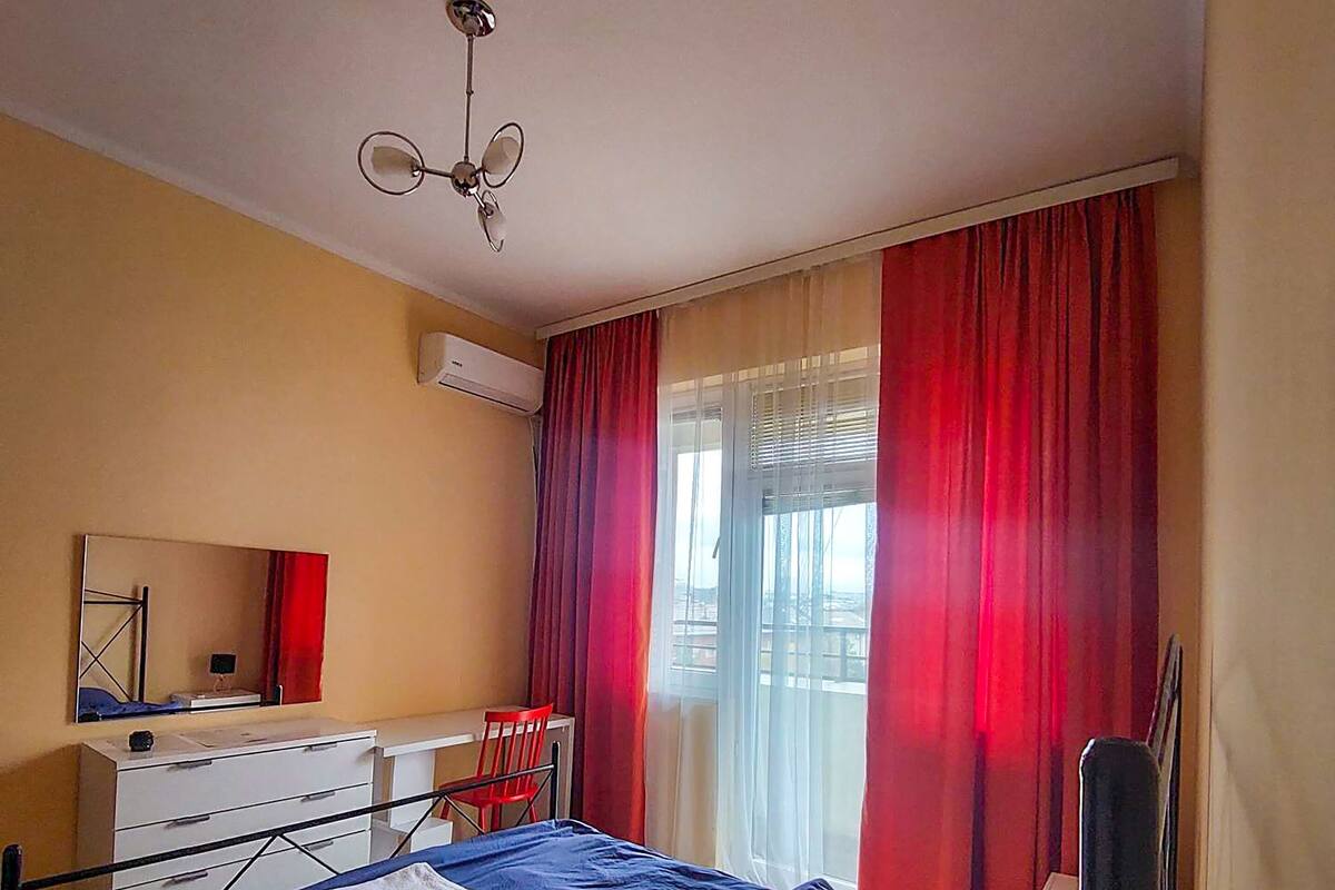 A bedroom with a bed, a desk, a chair, and a window with red curtains.
