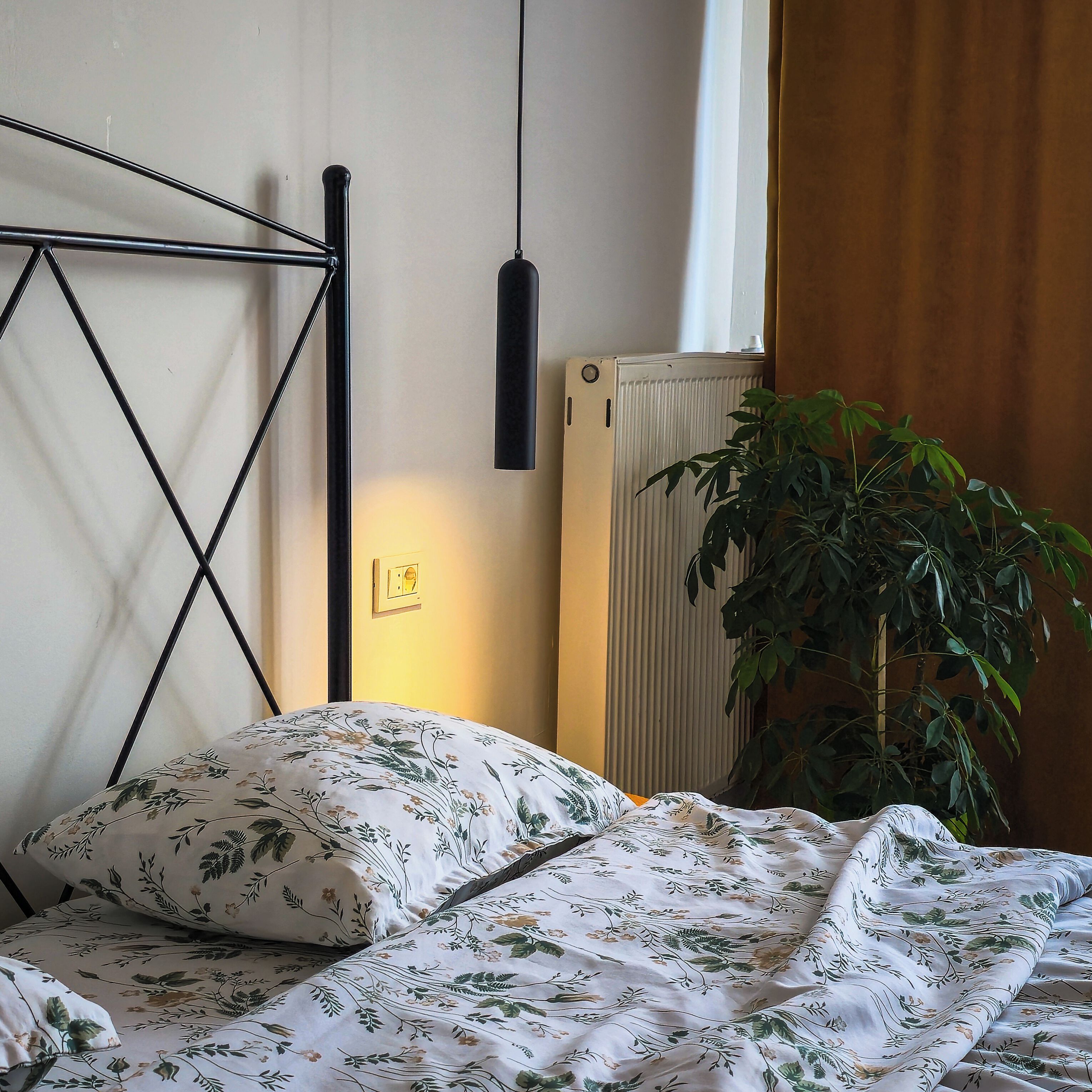 Sängyssä on kukallinen vuodevaatesetti, patteri ja ruukussa oleva kasvi.