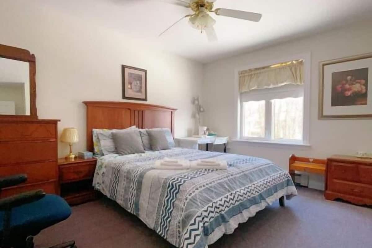 A bedroom with a bed, a desk, a chair, a dresser, a ceiling fan, and a window.