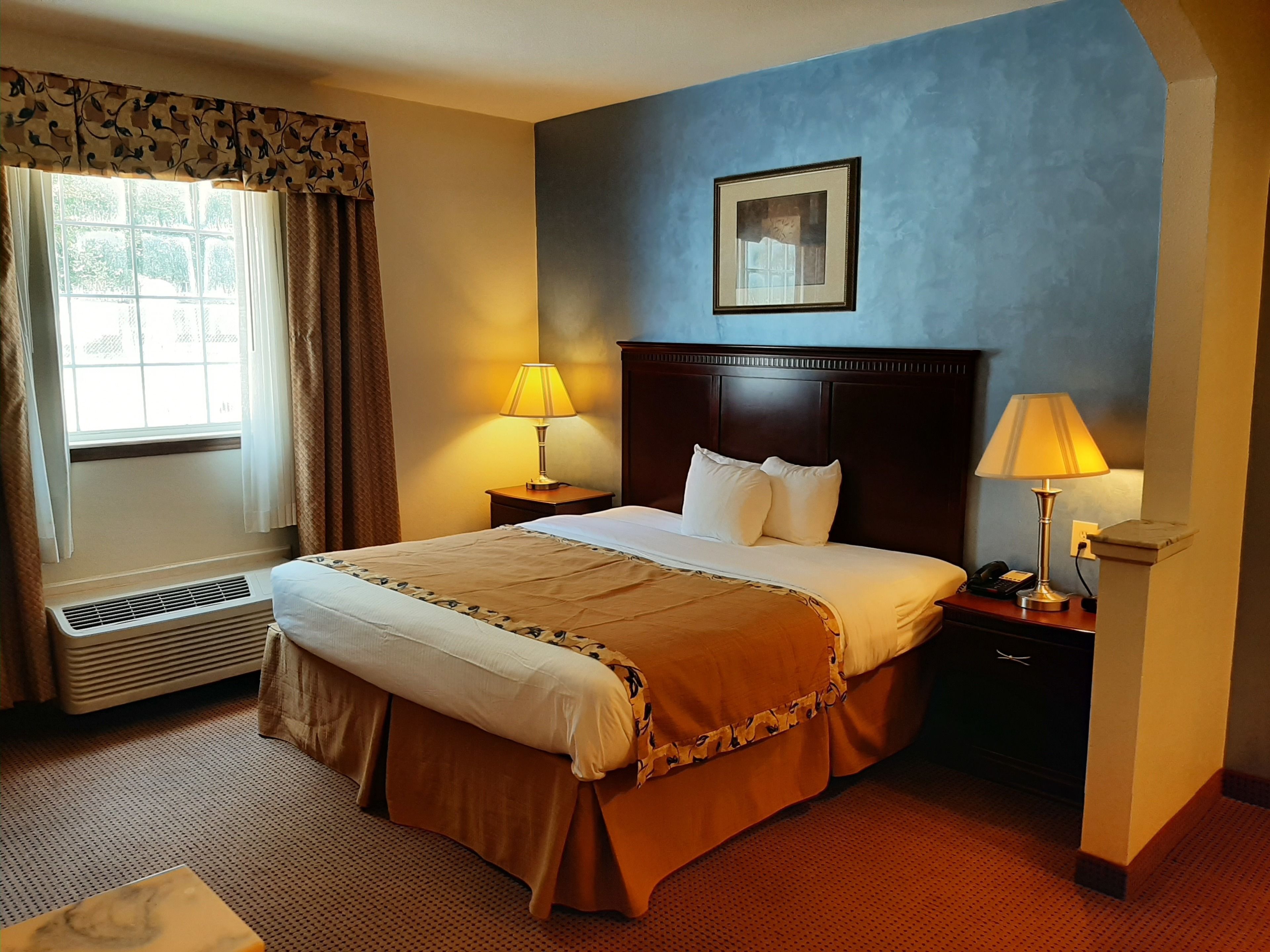 A hotel room with a large bed, two bedside lamps, a window with curtains, a framed picture on the wall, and a thermostat on the wall.