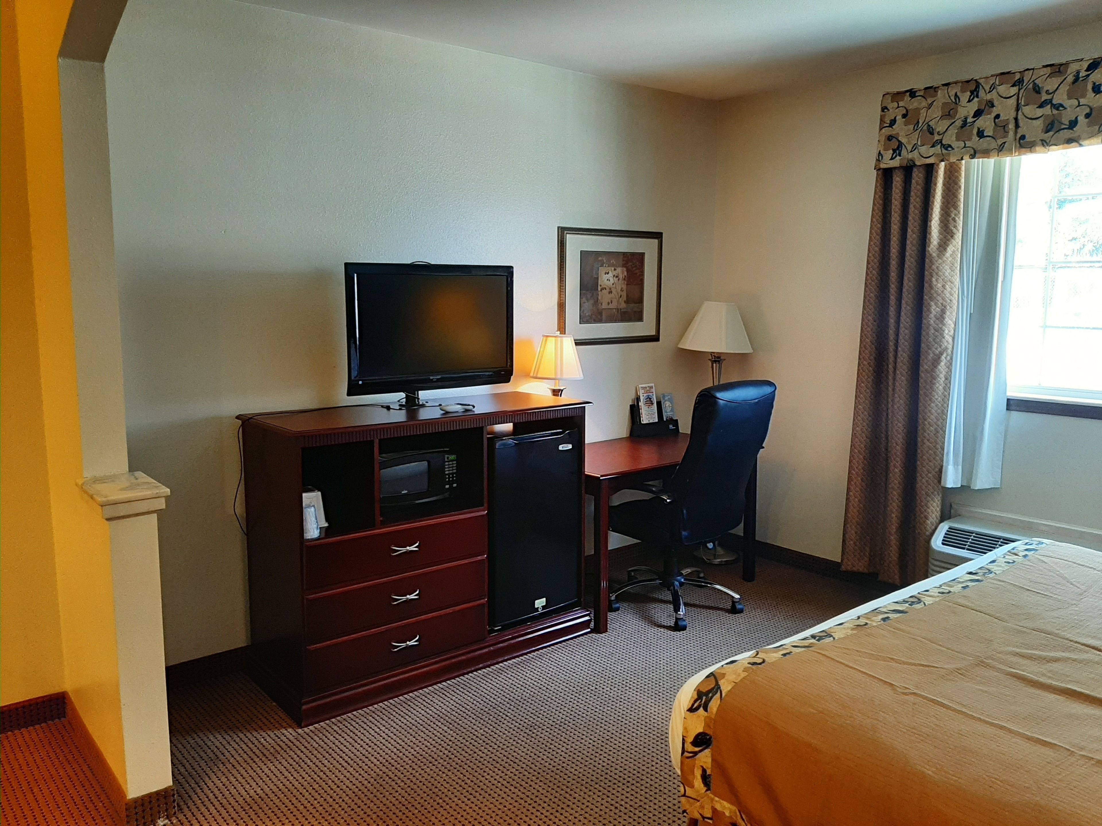 A hotel room with a bed, a desk with a television, a chair, a lamp, and a framed picture on the wall.