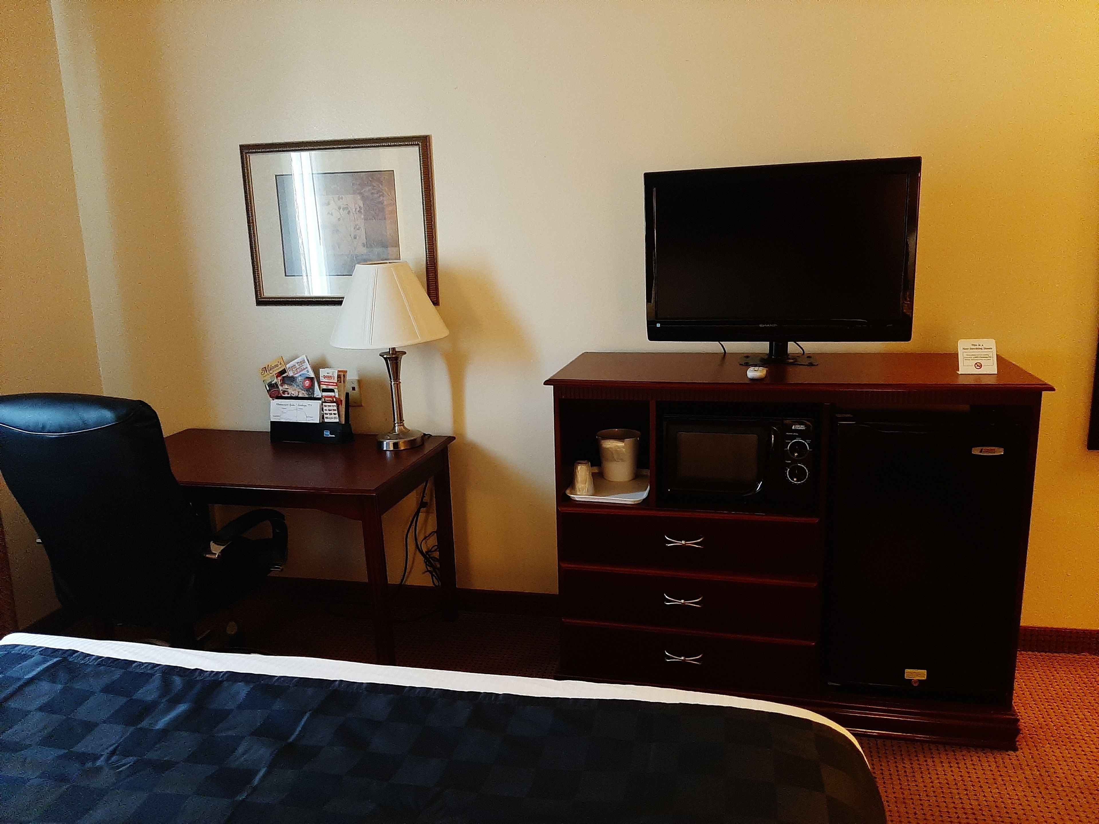 A hotel room with a bed, a desk with a lamp, a television, a microwave, and a dresser.