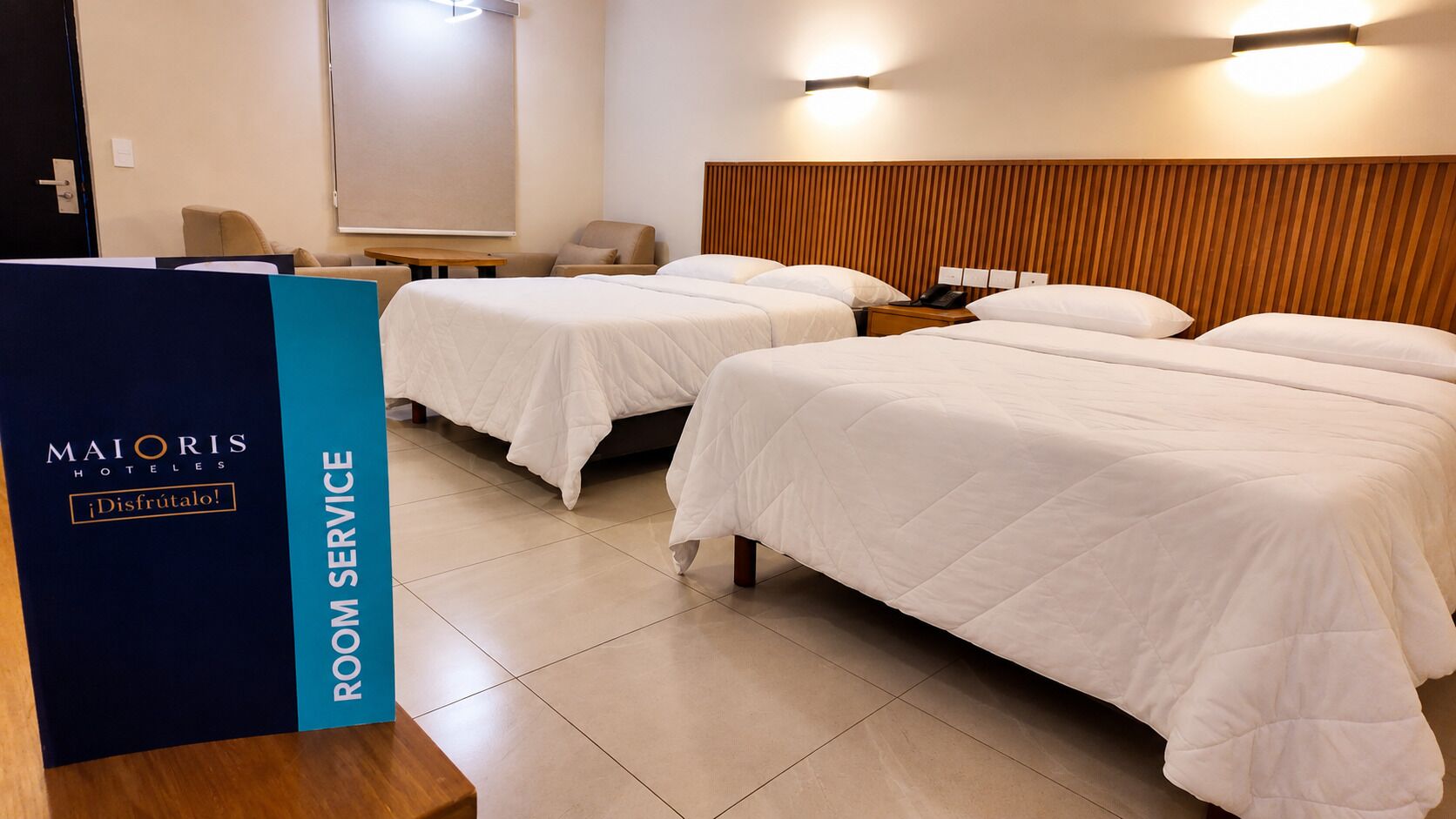 Camera d'albergo con due letti, una testiera in legno, un comodino con il telefono e una lampada fissata alla parete.
