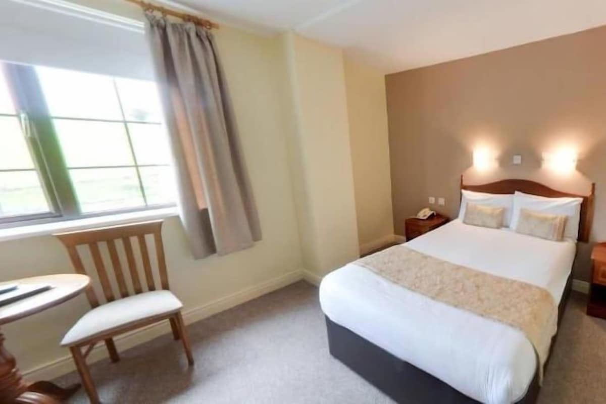 A hotel room with a bed, bedside table, nightstand, chair, and a window with curtains.