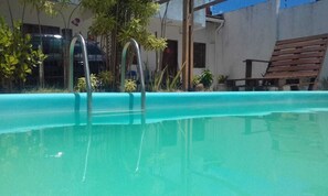 Piscina con escaleras de metal, rodeada de plantas y una silla de madera.