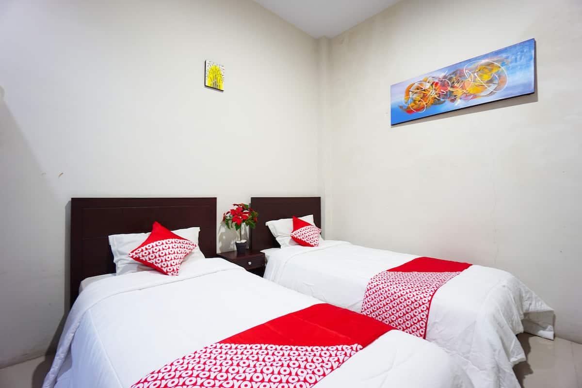 Quarto de hotel com duas camas, roupa de cama branca, detalhes vermelhos, um quadro na parede e um pequeno vaso com flores.