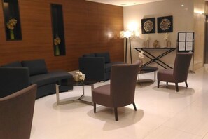 Un vestíbulo moderno con un sofá de color oscuro, una mesa redonda de centro y dos sillones. Hay una lámpara de pie y una obra de arte en la pared.