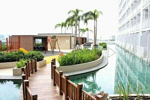 A pool area with a wooden deck, palm trees, and a building with balconies. - Breeze Residences by Ridged (Pasay)