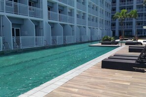 Zona de piscina de hotel con piscina cristalina, sillas de descanso y un edificio de varios pisos con balcones.
