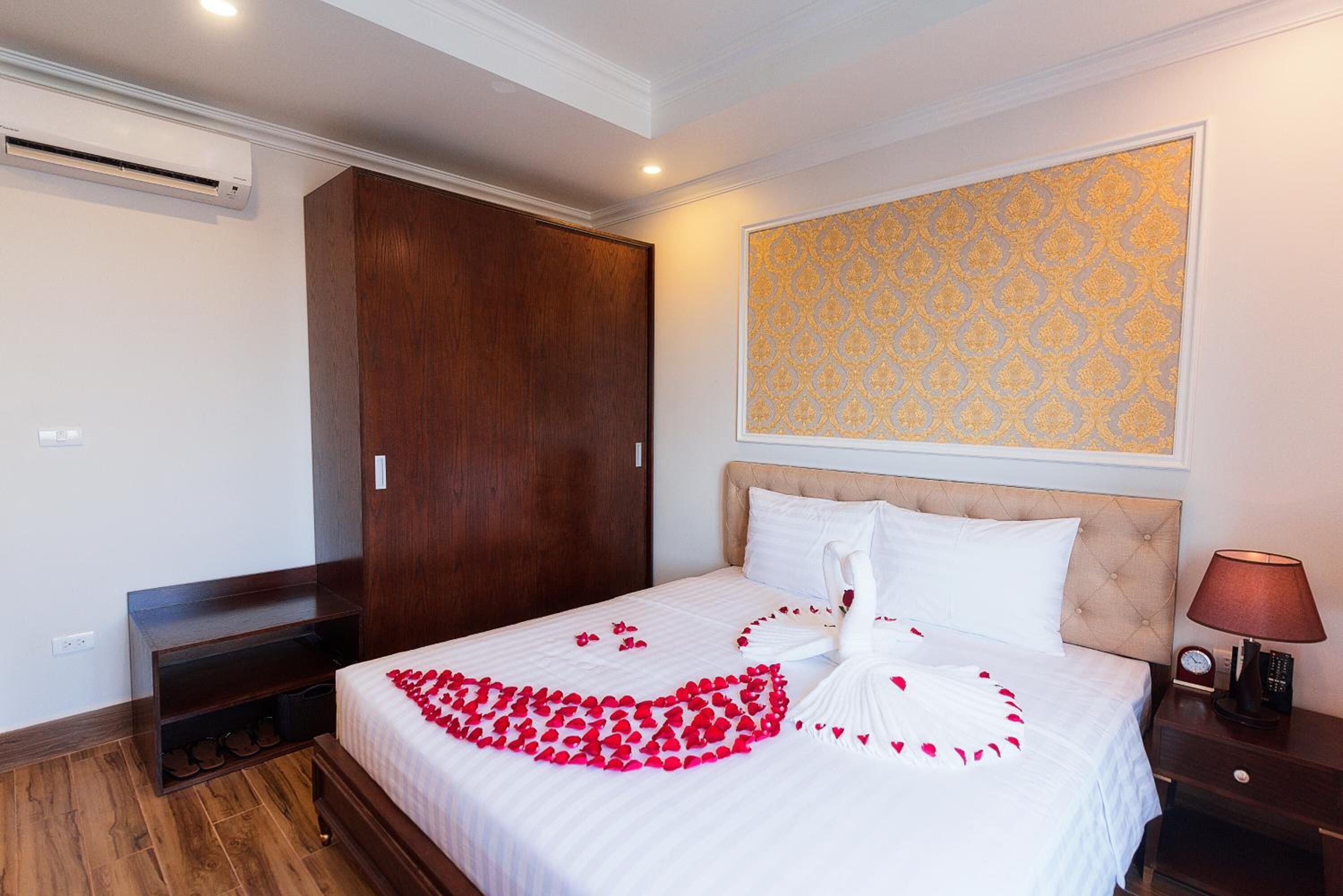 A hotel room with a bed, a wooden wardrobe, a wall-mounted air conditioner, a bedside table with a lamp, and a patterned wallpaper.