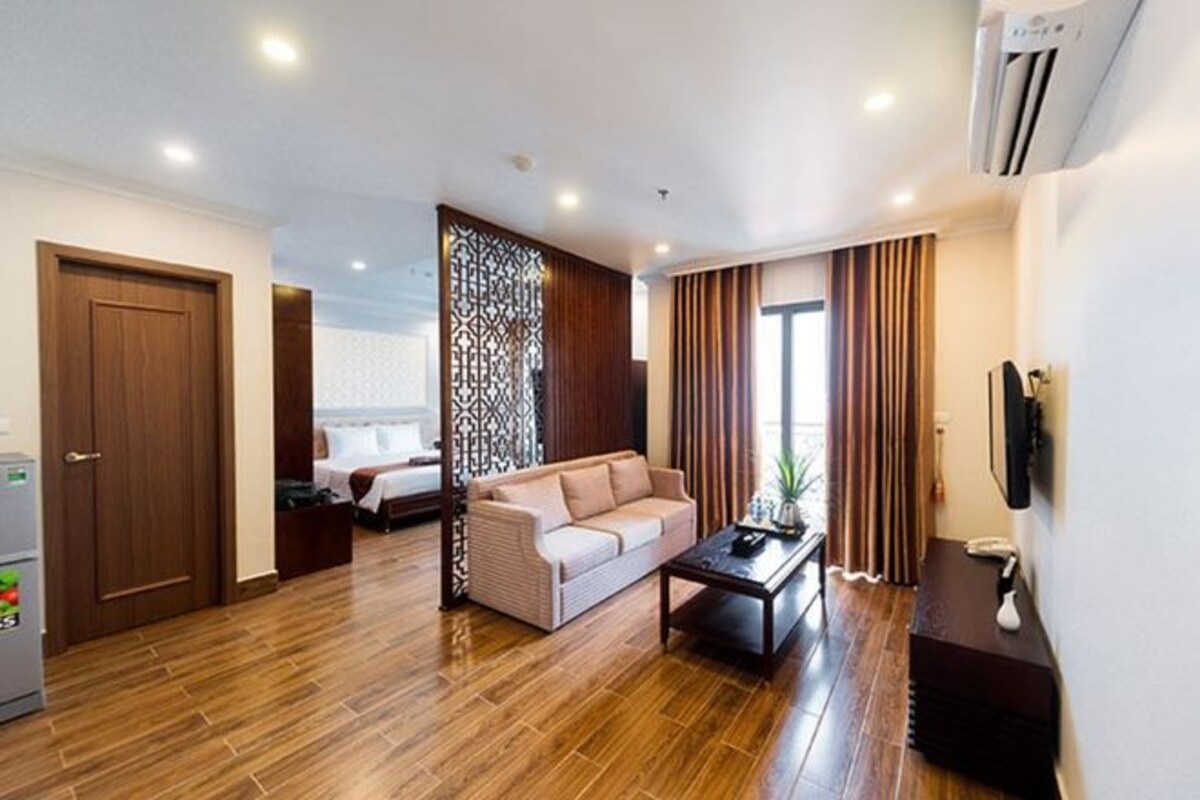 A modern hotel room with a wooden floor, a beige sofa, a coffee table, and a large window with curtains.