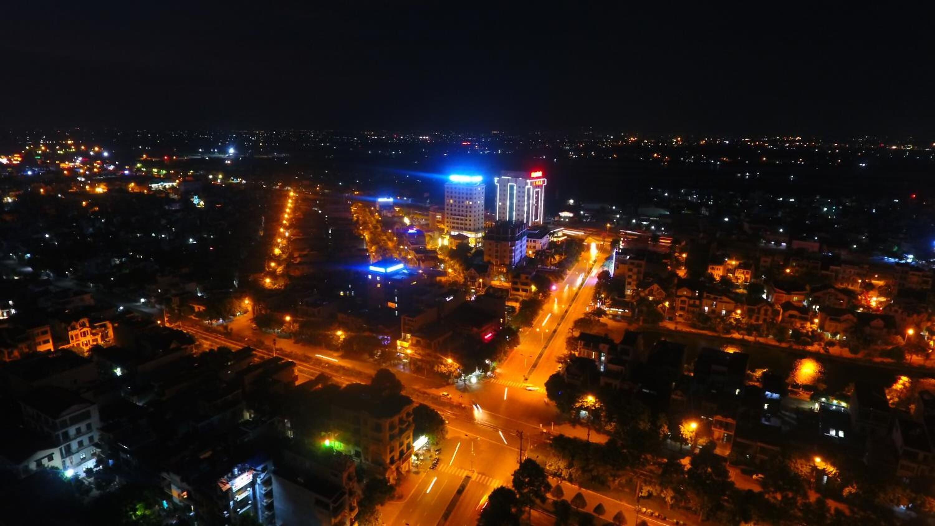 A nighttime aerial view of a cityscape with illuminated buildings, roads, and a prominent hotel with a lit sign.