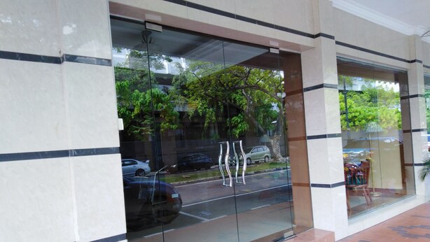 A modern building entrance with glass doors, a tiled floor, and a reflection of trees and parked cars. - Sara Hotel (Labuan)