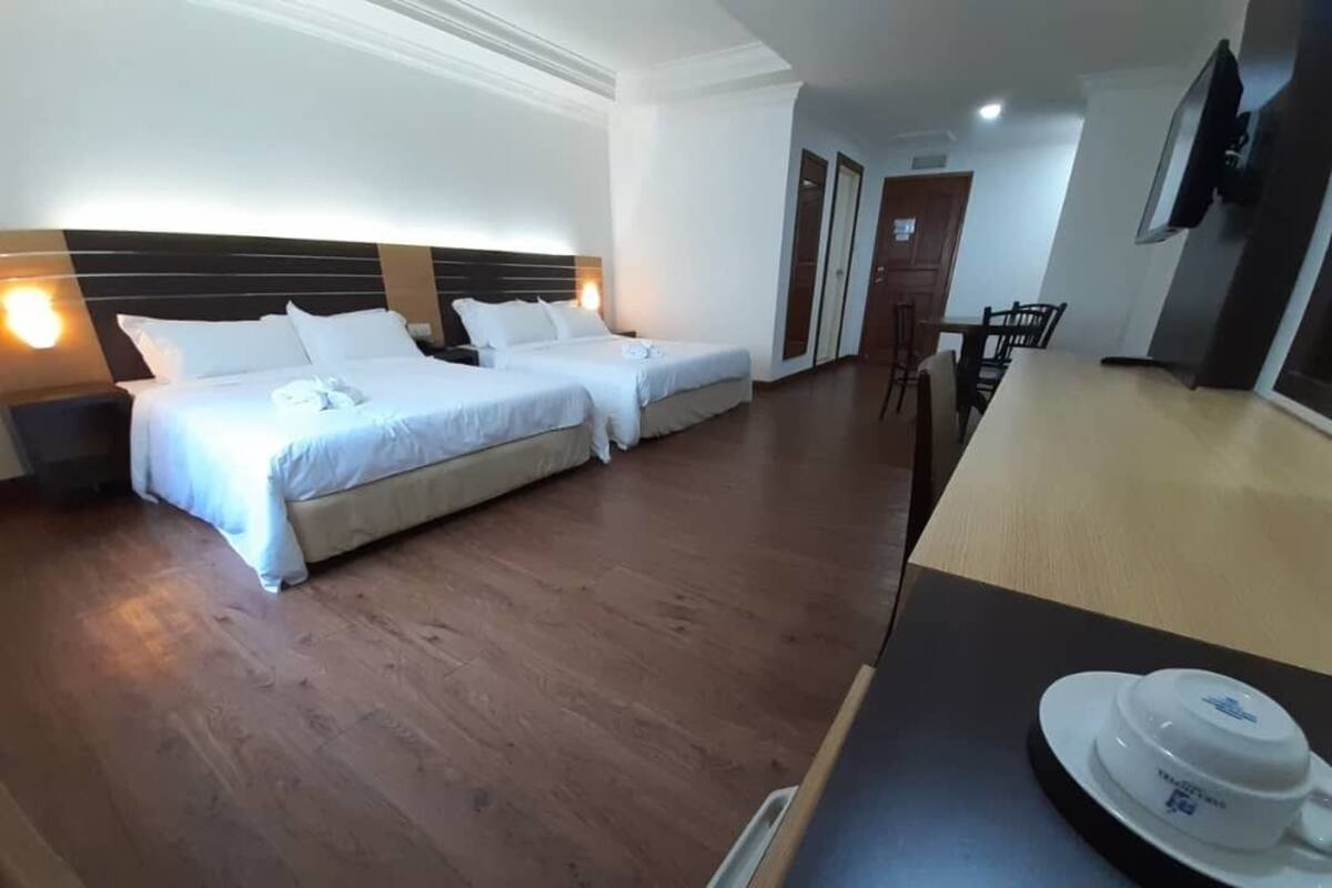 Habitación de hotel con dos camas individuales, piso de madera, una mesa de comedor con sillas y un televisor fijado a la pared.