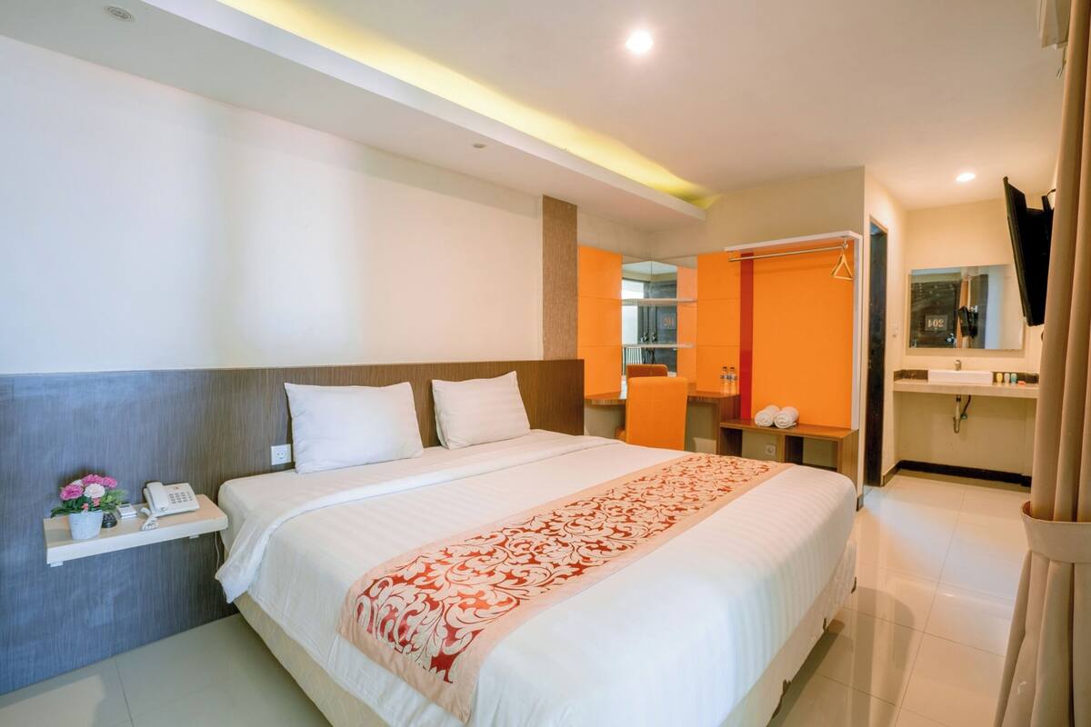 A hotel room with a large bed, a bedside table with a phone and flowers, an orange wall, a bathroom with a mirror and sink, and a TV mounted on the wall.