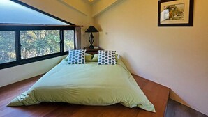 Ein Schlafzimmer mit großem Fenster, einem Bett mit gepunkteten Kissen, einem Nachttisch mit Lampe und einem gerahmten Bild an der Wand.